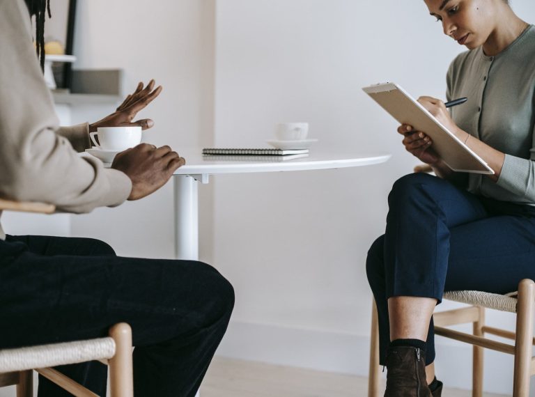 Crop ethnic female psychotherapist in formal clothes writing notes in clipboard while listening to black male patient complains in light studio